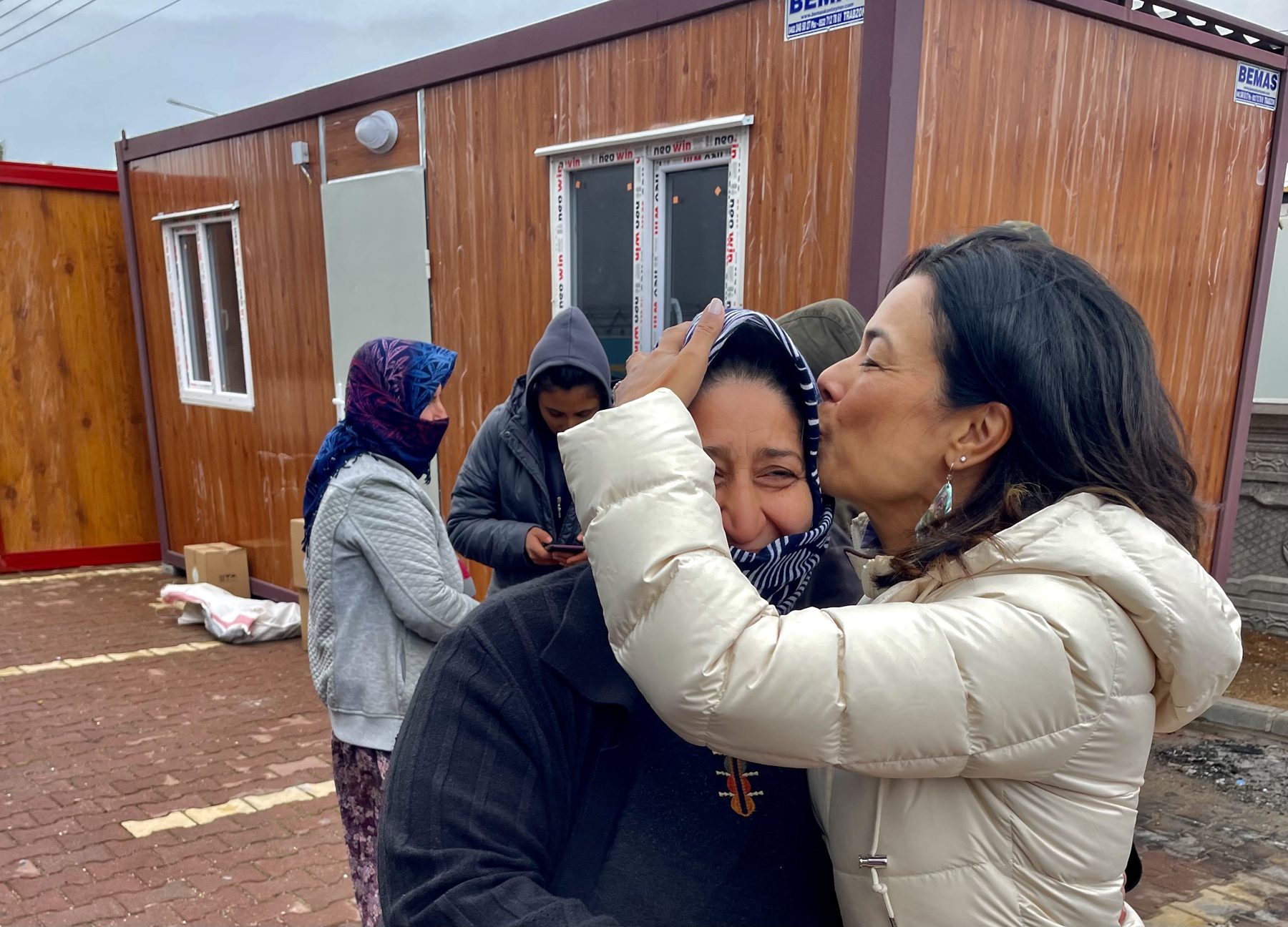 Charlene Nijmeh embraces a woman in a headscarf outside temporary container housing in Türkiye