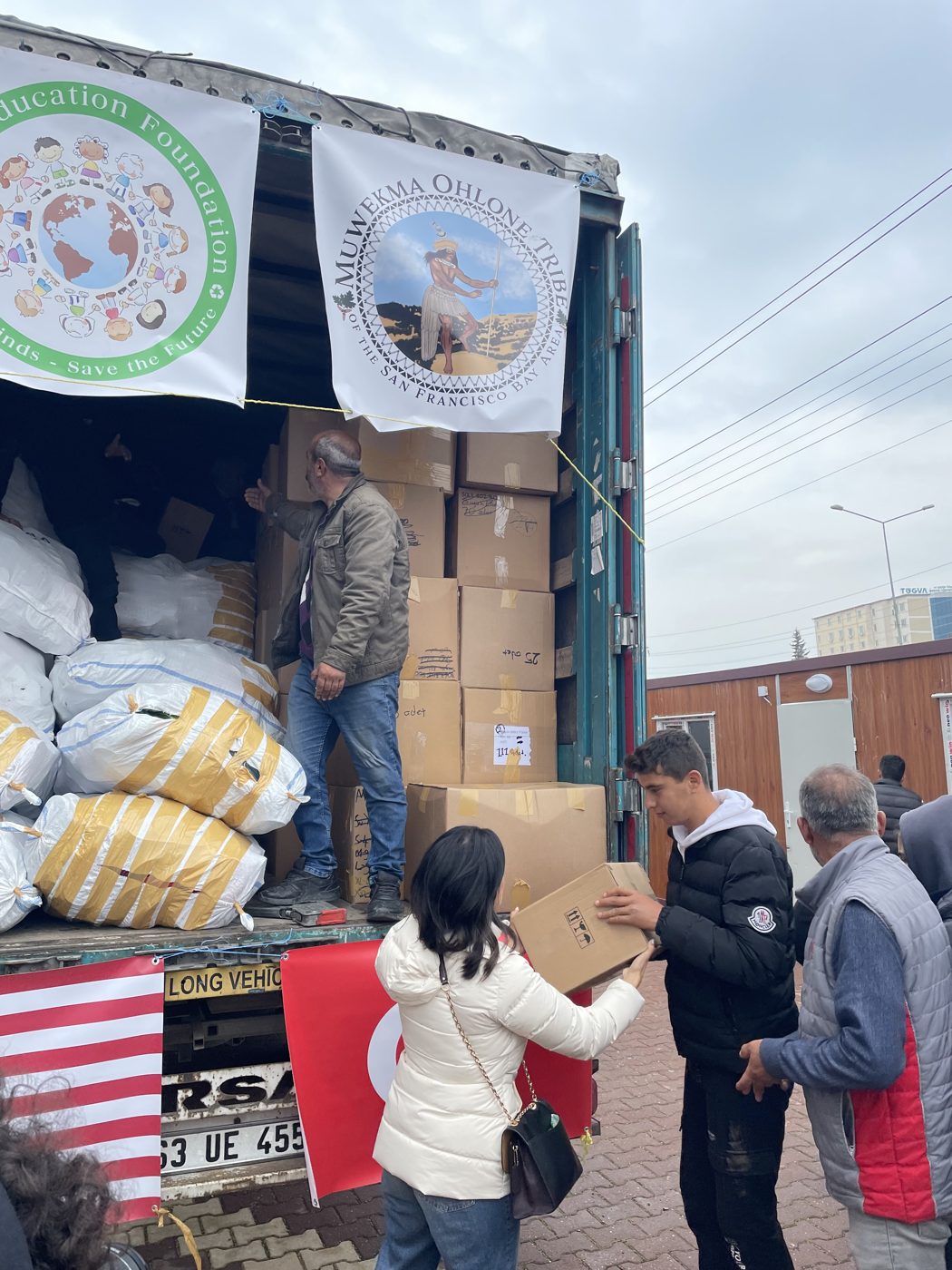 A relief truck loaded with boxes and sacks, hung with the Green Education Foundation seal and a Muwekma Ohlone Tribe banner, beside U.S. and Turkish flags